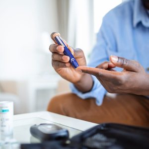 African man is sitting at the sofa at the home and taking blood from his finger due to diabetes. The daily life of a man of African-American ethnicity person with a chronic illness who is using glucose tester.