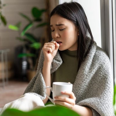 Sad ill asian girl staying on self quarantine during covid, catching flu and sitting at home with cup of tea near window, coughing in hand.
